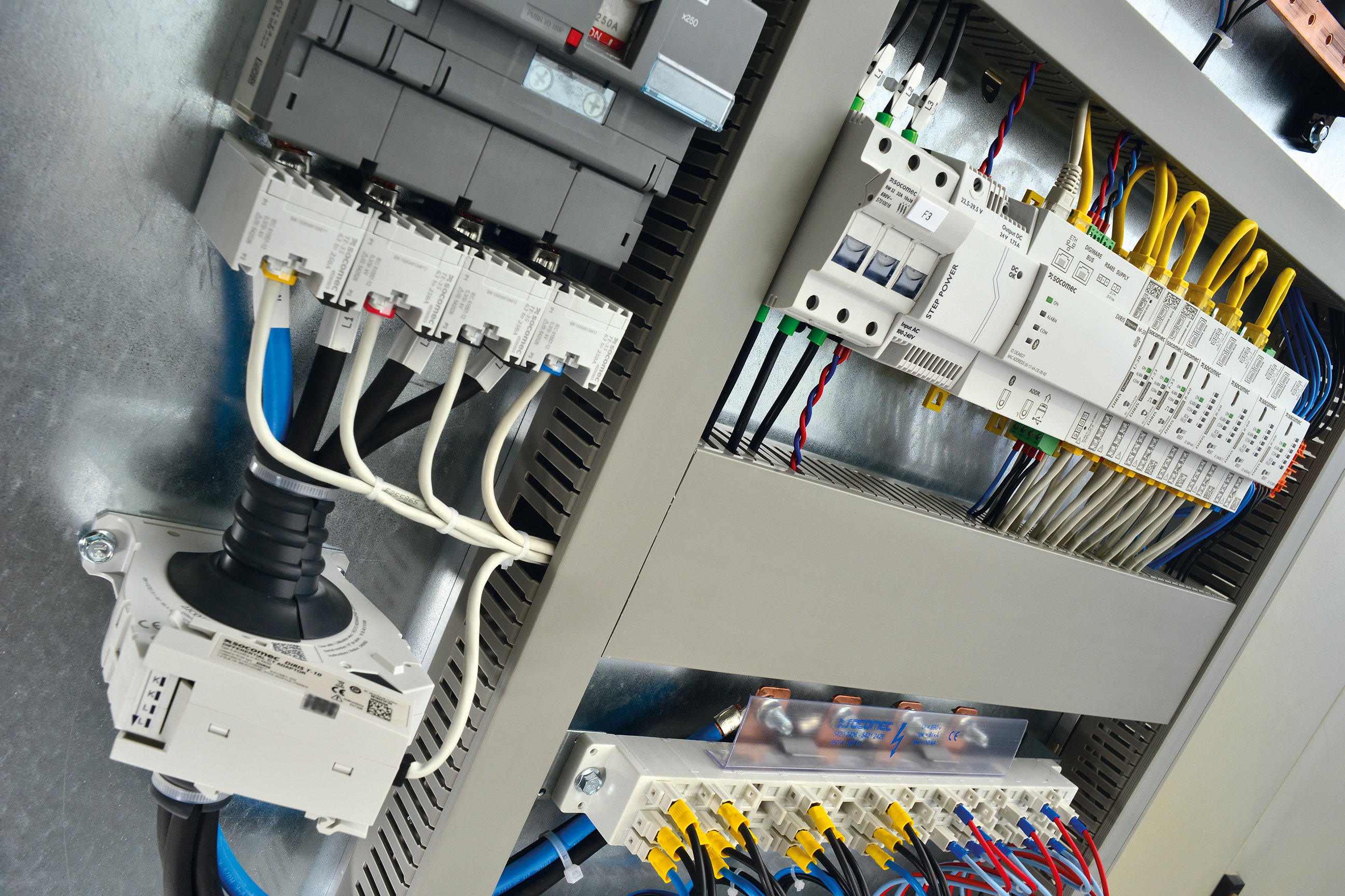Primo piano di un quadro elettrico aperto con componenti SOCOMEC: interruttori/dispositivi di protezione, moduli di comando e comunicazione, nonché morsettiere e blocchi di distribuzione cablati con conduttori di diversi colori.